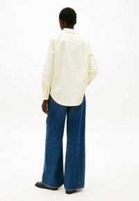 Camisa de manga larga amarilla clara con cuello puntiagudo, de corte holgado y mangas con puños, combinada con jeans de pierna ancha de talle alto en azul.