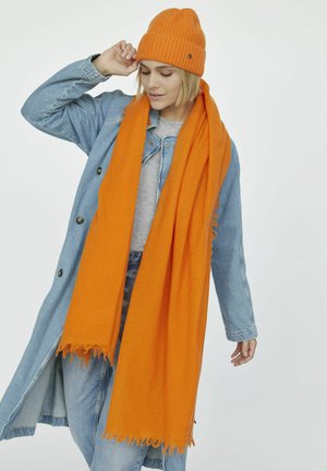 Helle orange Strickmütze und Schal, kombiniert mit einer Jeansjacke über einem grauen Oberteil. Der Schal hat fransige Kanten für zusätzlichen Textur.
