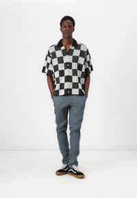 Chemise à carreaux noir et blanc à manches courtes avec un col, associée à un pantalon gris. Le modèle porte des baskets noir et blanc avec des semelles en gomme.