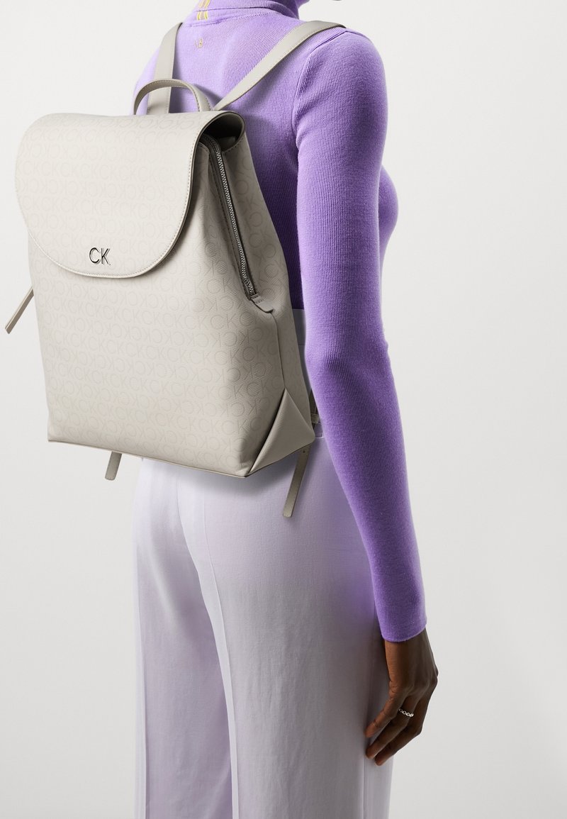 Jasnoszary skórzany plecak o gładkim wykończeniu z wytłoczonym logo CK. Posiada zaokrągloną klapę oraz regulowane paski.