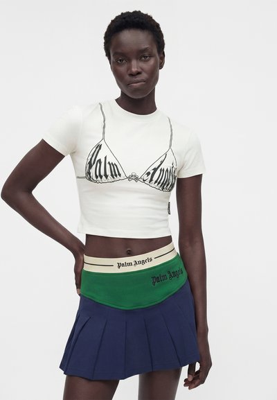 Το μοντέλο φοράει λευκό κροπ top με γραφικό σχέδιο, πράσινη και κρεμ ζώνη με το κείμενο "Palm Angels" και μια σκούρα μπλε πλισέ φούστα.