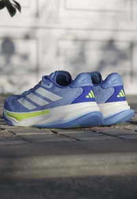 Blue athletic shoes with a textured upper, three white stripes, a green accent, and a white, ridged sole. Notable heel and toe design.