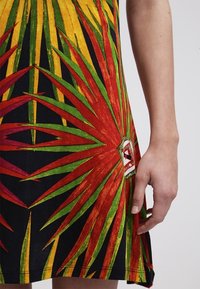 Gros plan sur une main reposant à côté d'une robe colorée arborant un motif géométrique étoilé audacieux en rouge, vert et jaune sur un fond noir.