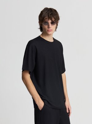 Giovane uomo con occhiali da sole rotondi, una camicia nera texture larga a maniche corte e pantaloni neri, in piedi davanti a uno sfondo grigio semplice.