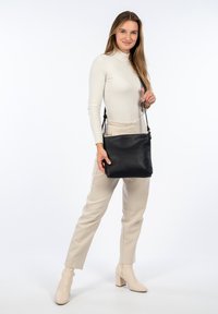 Borsa a tracolla in pelle nera con superficie texturizzata, forma rettangolare, tracolla regolabile e branding sottile. Il modello indossa pantaloni beige e stivali.