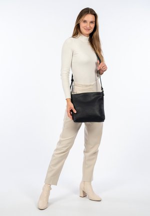 Borsa a tracolla in pelle nera con superficie texturizzata, forma rettangolare, tracolla regolabile e branding sottile. Il modello indossa pantaloni beige e stivali.