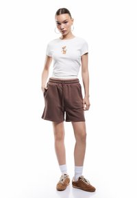 Camiseta blanca de algodón con gráfico de un oso, combinada con pantalones cortos de jersey marrones. El modelo lleva calcetines blancos hasta la altura del tobillo y deportivas marrones con cordones.