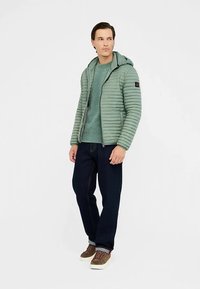 Groene gewatteerde jas met horizontale stiksels, capuchon en ritssluiting, gedragen over een getextureerde teal trui. Gecombineerd met donkere jeans en bruine schoenen.