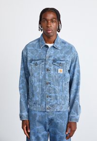 Hellblaue Jeansjacke mit floralem Camouflage-Muster, ausgestattet mit Knopfverschluss, zwei Brusttaschen und einem Markenzeichen auf der linken Tasche.