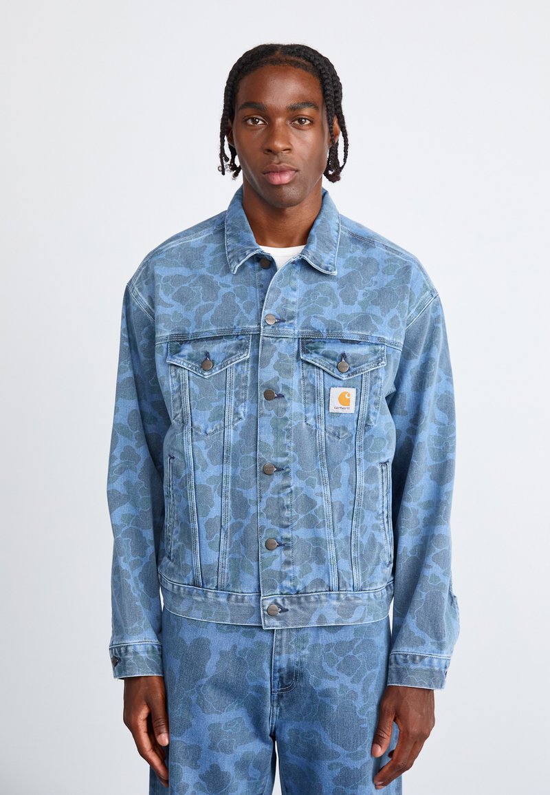 Hellblaue Jeansjacke mit floralem Camouflage-Muster, ausgestattet mit Knopfverschluss, zwei Brusttaschen und einem Markenzeichen auf der linken Tasche.