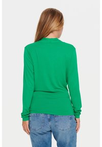 Maglia verde a maniche lunghe realizzata in tessuto morbido, con un design aderente e colletto alto. Abbinata a jeans in denim blu chiaro.