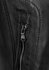 Sort læderjakke med et nærbillede af en lynlås med en blank trækflap, omgivet af synlige sømme og en tekstureret overflade.