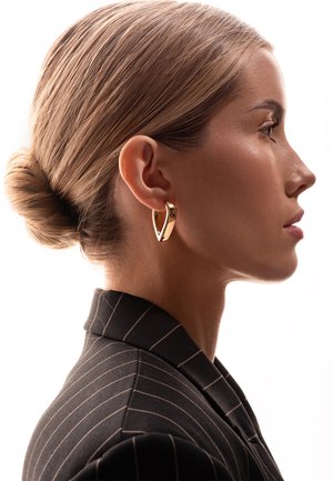 Złota kolczyk w kształcie obręczy o unikalnym wzorze w kształcie V, z polerowanym wykończeniem, noszony przez modelkę z włosami w schludnym kokiem i w marynarce w paski.