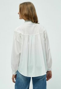 Camicia bianca a maniche lunghe con yoke arricciato sul retro, colletto alto e polsini con bottoni, abbinata a jeans blu dalla vestibilità rilassata.