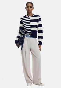 Gestreepte trui in marineblauw en crèmekleurig, met een uniek asymmetrisch ontwerp; gecombineerd met brede crèmekleurige broek en witte schoenen.
