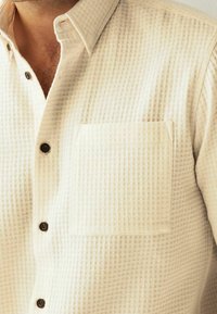 Chemise boutonnée couleur crème avec un motif tissé texturé. Elle présente une poche poitrine et des boutons marron pour la fermeture.