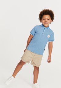 Lichtblauwe polo met een gewoven ontwerp, witte kraag en mouwafwerkingen, geborduurd op de borst; gecombineerd met beige opgerolde shorts en witte schoenen.