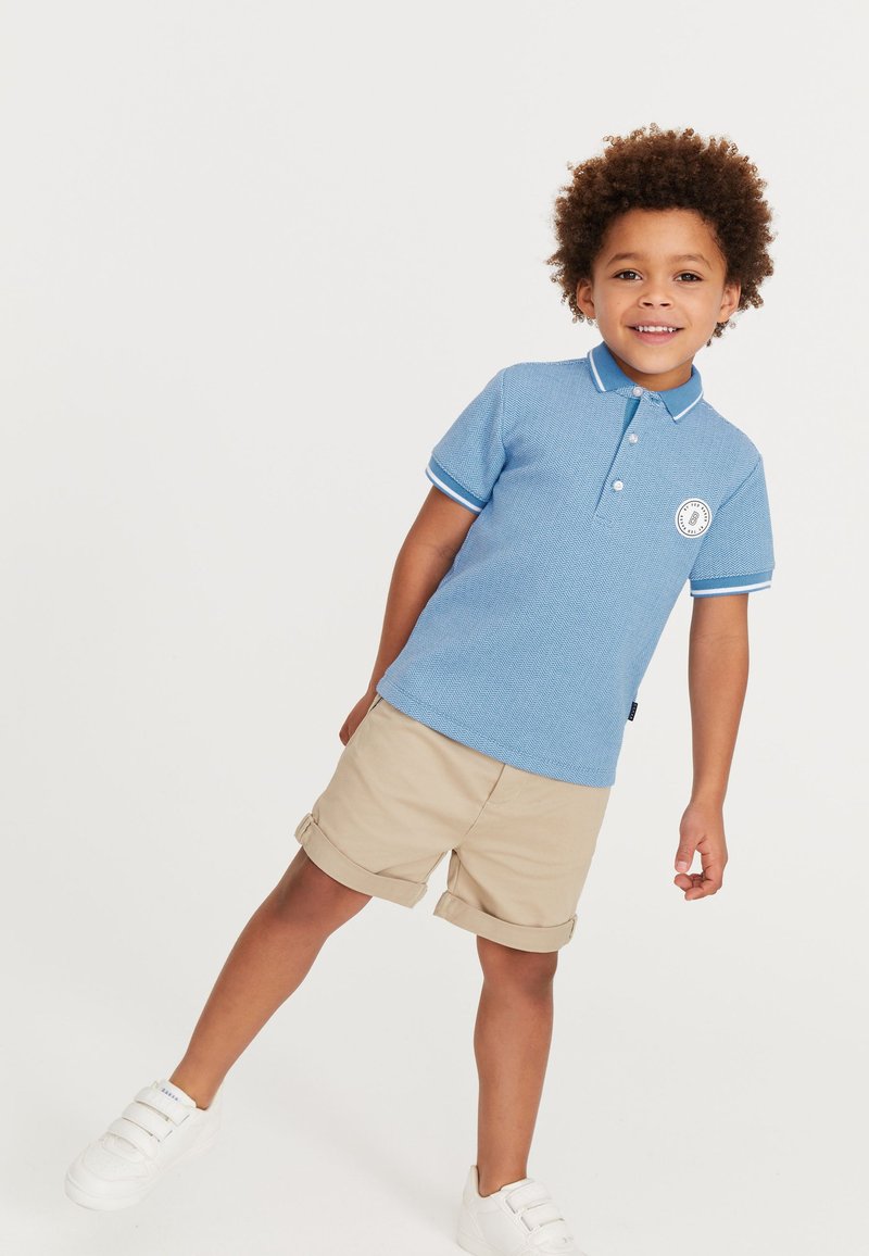 Lichtblauwe polo met een gewoven ontwerp, witte kraag en mouwafwerkingen, geborduurd op de borst; gecombineerd met beige opgerolde shorts en witte schoenen.
