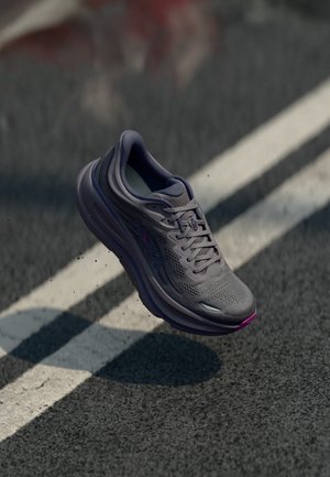 Chaussure de course grise avec des accents violets en plein vol au-dessus d'une route en asphalte avec des marquages blancs.