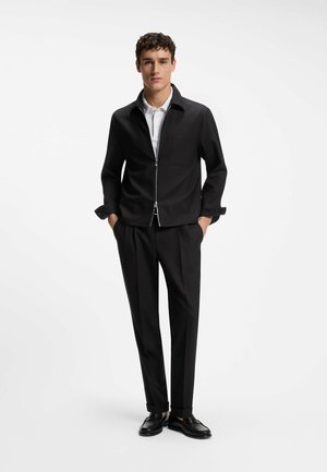 Young man wearing black zip-up jacket, black tailored pants, white polo shirt, and black loafers, standing with hands in pockets.