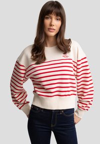 Sudadera crema con franjas horizontales rojas, puños y dobladillo acanalados, con logo bordado en el pecho. Vaqueros oscuros con textura de denim.