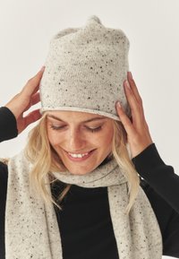 Lichtgrijze gebreide muts met zwarte spikkels, aansluitende vorm; gecombineerd met een bijpassende lichtgrijze sjaal. De textuur ziet er zacht en gezellig uit.