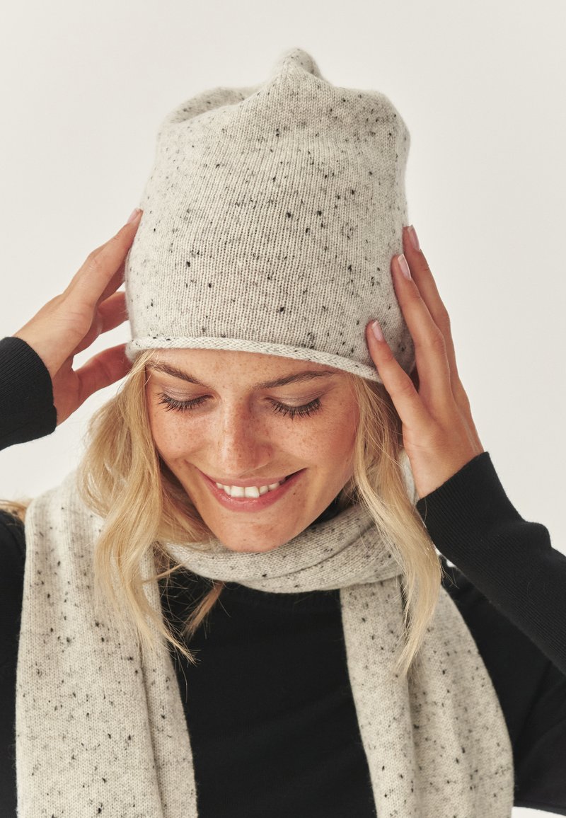 Lichtgrijze gebreide muts met zwarte spikkels, aansluitende vorm; gecombineerd met een bijpassende lichtgrijze sjaal. De textuur ziet er zacht en gezellig uit.