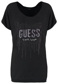 T-shirt noir à manches courtes en tissu doux, arborant "GUESS" en lettres larges ornées de sequins argentés et un design décontracté.