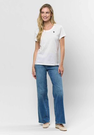 Bela kratka majica s kratkimi rokavi in okroglo ovratnikom ter majhnim logotipom, v kompletu s svetlo modrimi širokimi hlačami in bež natikači.