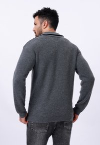 Grau gerippter Strickpullover mit Schalkragen, langen Ärmeln und lockerer Passform; kombiniert mit dunklen Jeans, die Detailaufnahmen des strukturierten Stoffs zeigen.