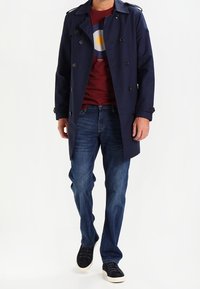 Marinblå trenchcoat med dubbelknäppt design, axelremen, tillsammans med en vinröd grafisk t-shirt och blå jeans. Svarta sneakers fulländar looken.