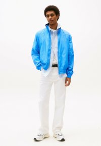 Lichtblauwe ritsjack met elastische manchetten, gecombineerd met een gestreept shirt, witte broek en lichte sneakers, wat zorgt voor een casual outfit.