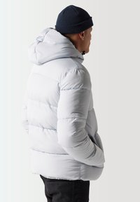 Chaqueta de plumas azul claro con capucha, textura acolchada y puños elásticos. Combinada con un gorro de punto azul marino. Se muestra desde la parte de atrás.