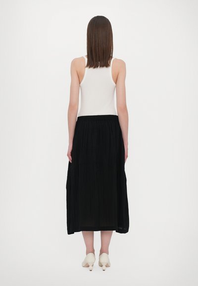 Sort plisseret nederdel med elastisk talje, sammen med en hvid tanktop og cremegule hæle. Bagudblik af outfittet.