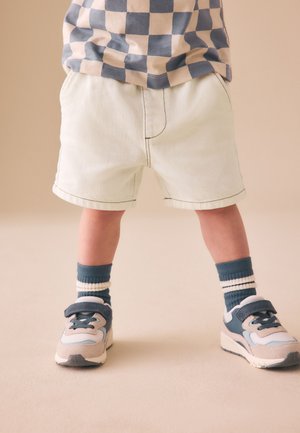 Småbarn iført beige shorts, blå og beige sneakers med velcrolukning, blå og beige stripede sokker og en blå og beige ternet skjorte.