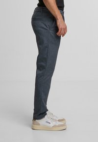 Pantaloni grigio scuro con texture e vestibilità slim, caratterizzati da un davanti piatto e tasche laterali, abbinati a sneaker beige e bianche.
