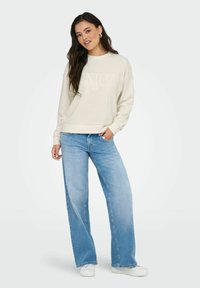 Jeune femme se tient debout portant un sweat-shirt crème avec le texte "ENJOY", un jean large bleu clair, des baskets blanches et des boucles d'oreilles en argent sur fond uni.