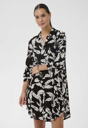 Kobieta ubrana w czarno-białą sukienkę na guziki w kwiaty z rękawami do połowy łokcia, pozująca z delikatnym uśmiechem i skrzyżowanymi na talii rękami.