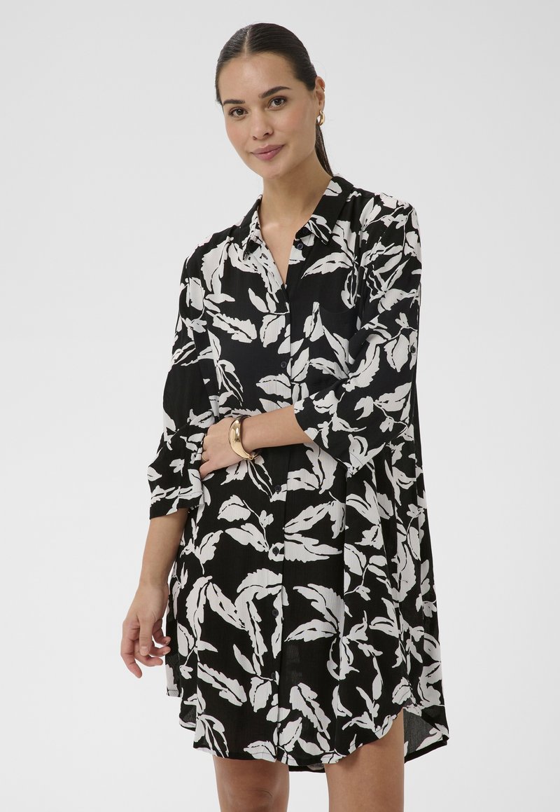 Femme portant une robe boutonnée à fleurs noire et blanche aux manches trois-quarts, posant avec un léger sourire et les mains croisées à la taille.