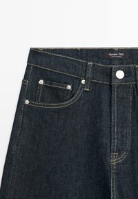 Jeans in denim scuro con un design classico, chiusura con bottone, cuciture in rame, dettagli sulle tasche frontali e etichetta del marchio all'interno.