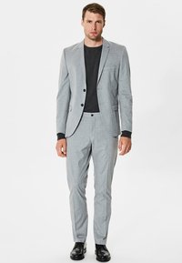 Traje a medida de color gris claro con solapas afiladas, diseño de un solo botón y bolsillos frontales. Una camisa negra debajo y zapatos negros completan el conjunto.