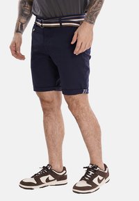 Pantaloni corti blu realizzati in cotone, con risvolti, tasche frontali e una cintura a righe. Sneakers marroni e bianche completano l'outfit.