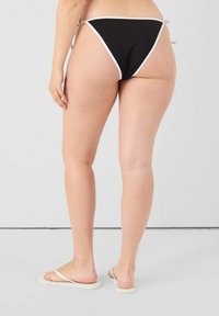 Bas de bikini noir avec bord blanc, présentant un design minimaliste et des liens latéraux réglables. Tissu lisse, coupe taille basse.