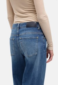 Niebieskie dżinsy z denimu o luźnym kroju, wyposażone w dwie tylne kieszenie, skórzaną metkę na pasku oraz subtelne przeszycia.