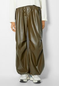Olivgrön imitation läderbyxor med elastiskt midjeband, avslappnad passform och subtil lyster, kombinerat med vita och flerfärgade sneakers.