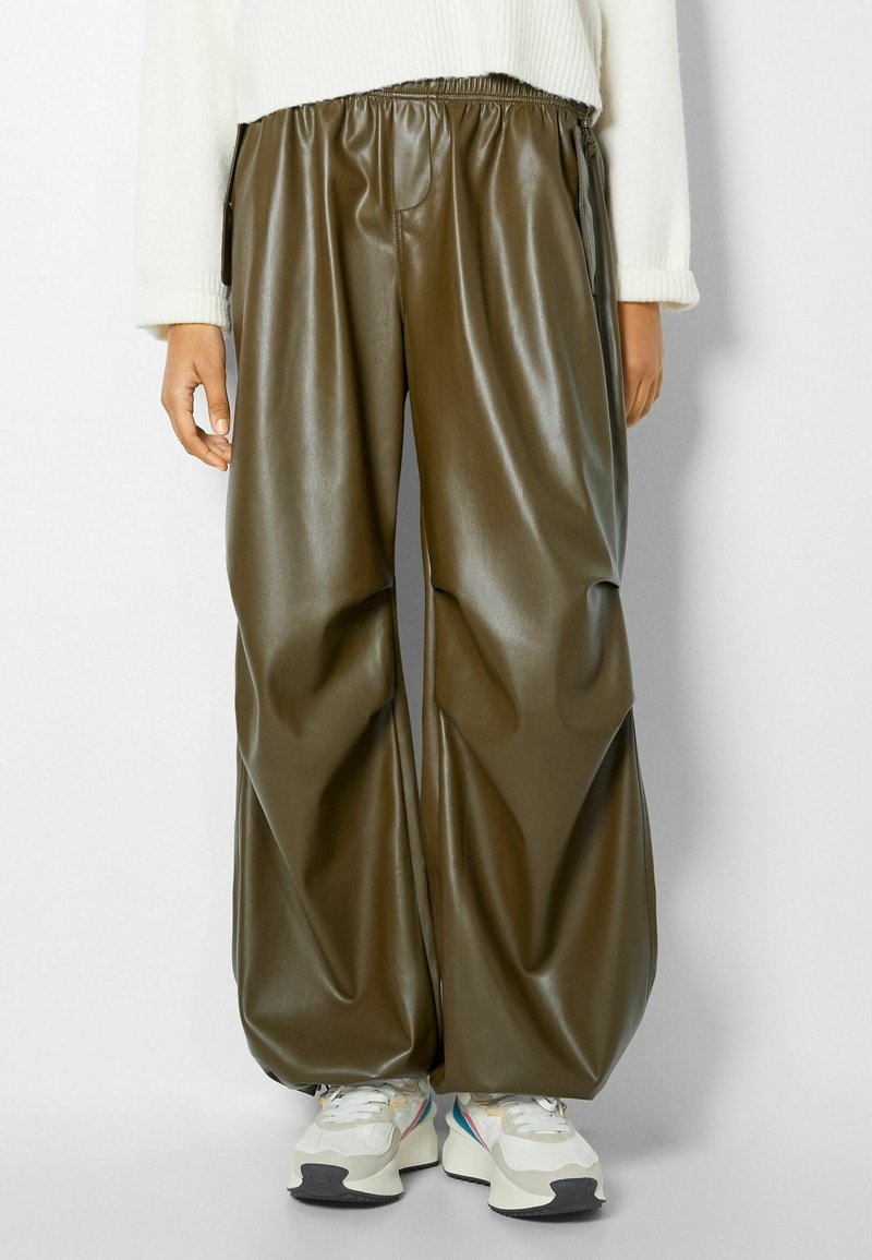 Olivgrön imitation läderbyxor med elastiskt midjeband, avslappnad passform och subtil lyster, kombinerat med vita och flerfärgade sneakers.