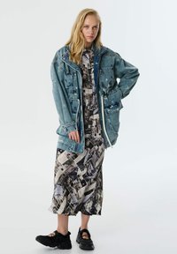 Blonde vrouw in een oversized blauwe spijkerjack over een lange patronenjurk en zwarte chunky schoenen, staand tegen een effen achtergrond.