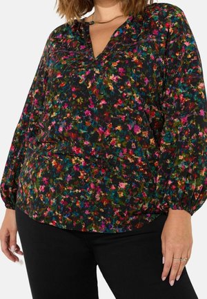 Mujer con blusa negra de manga larga con un colorido patrón floral y pantalones negros, mostrando el torso y las manos.