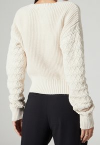 Licht crèmekleurige gebreide trui met een geribde ronde halslijn, gestructureerde mouwen met een geometrisch patroon, en een korte geribde zoom.