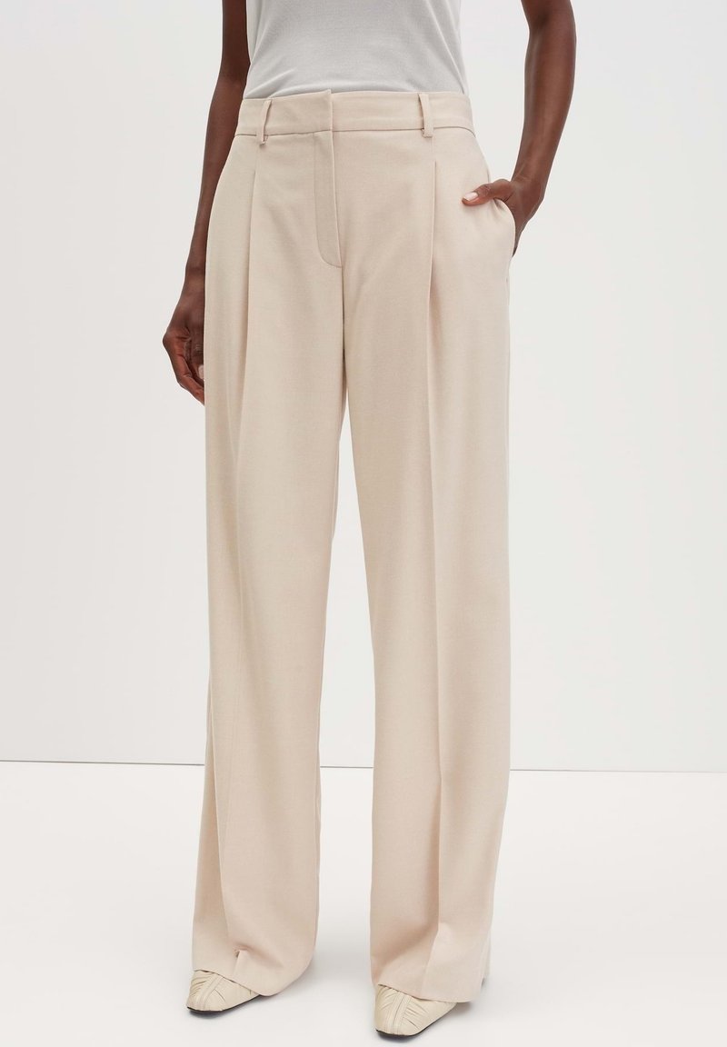 Pantalon beige à jambes larges avec des plis porté par une personne en haut blanc, montrant des poches latérales et une coupe taille haute.
